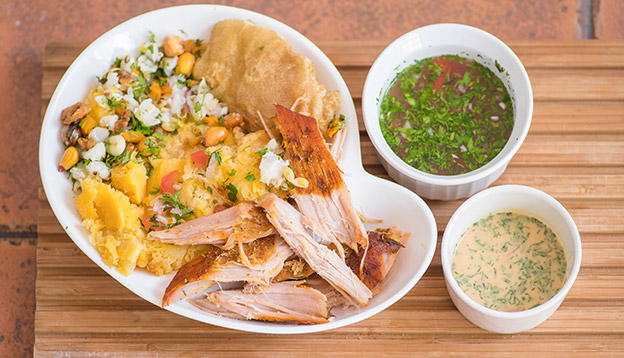 Above view of delicous hornado with mashed potatoes with salad, tostado and mote inside of a white plate with two smalls bowls with green and pink sauce